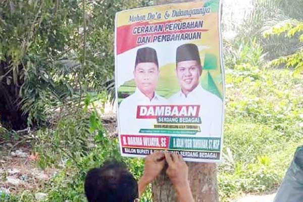 SPANDUK: Orang suruhan oknum Kades (DS), berinisial (R) memasang spanduk Balon Bupati dan Wakil balon Bupati Sergai yang dipajang di pohon, Selasa (23/6).